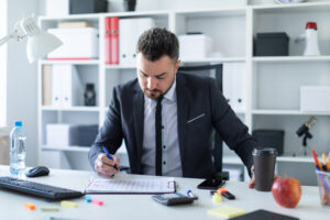 A man is sitting in the office at the table, holding a glass of coffee and a pen in his hand and working with documents